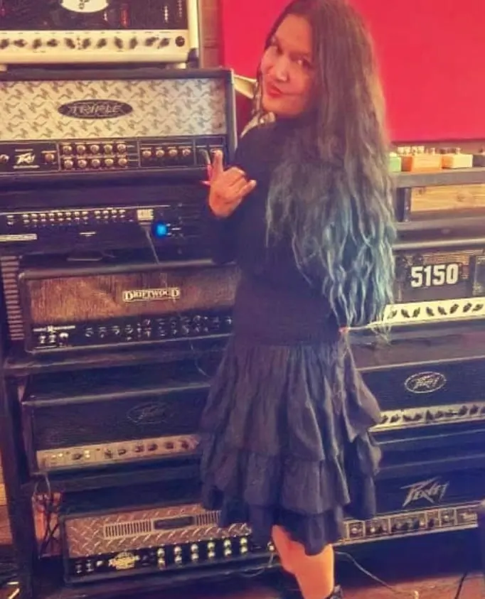 A figure with long blue-tinted hair throws devil horns in front of a stack of Peavey, 5150 and Driftwood amplifier heads.