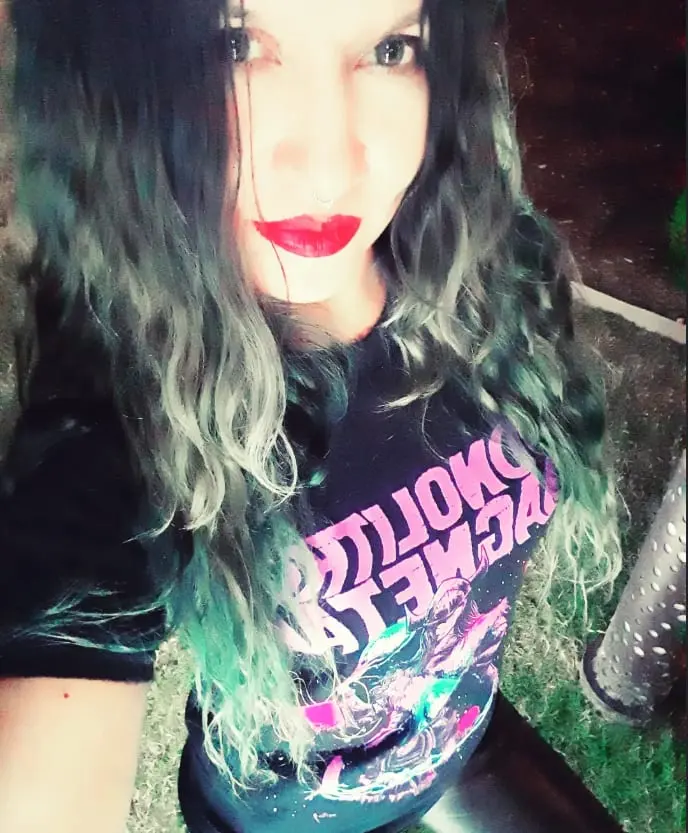 A person with teal and silver hair wearing a Monolithic Magnetar shirt, photographed outdoors at night.