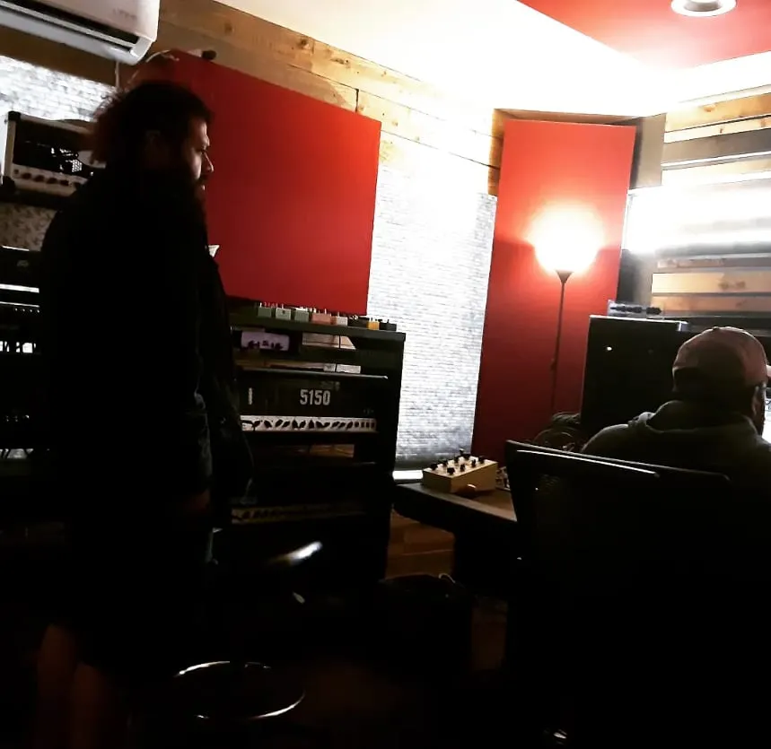 Two people in a control room lit red, a 5150 amplifier head and outboard racks along the wall.