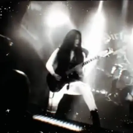 A black and white live shot of a guitarist mid-song, backlit by stage lights with the drum kit behind them.