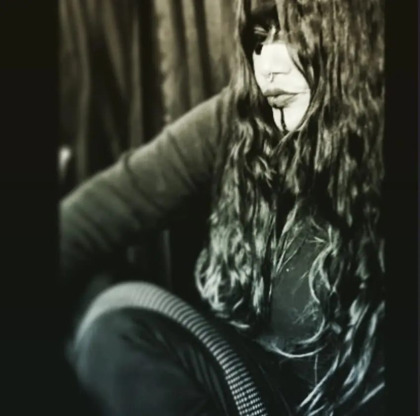A high-contrast black and white portrait of a long-haired figure seated, face half hidden behind their hair.