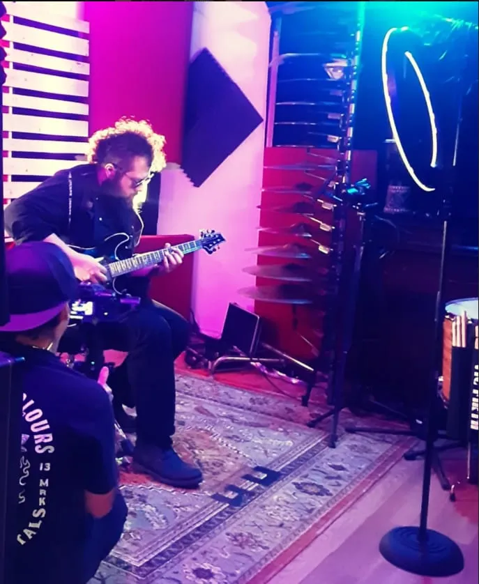 A guitarist plays seated under magenta and teal stage lights while a camera operator films from the floor.