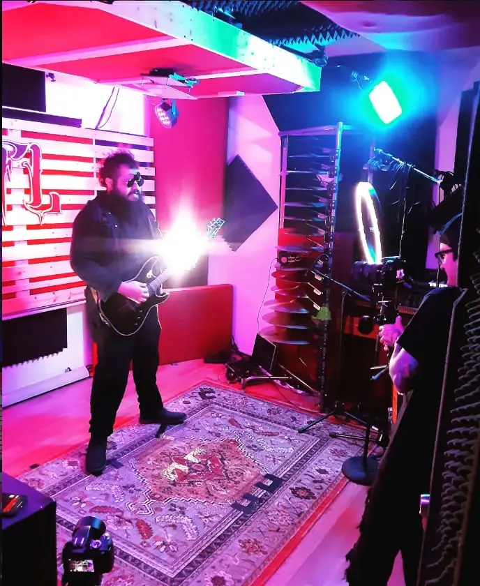 A wide view of the video shoot: a guitarist playing under coloured lights on a rug, ring light and camera rigs framing the room.