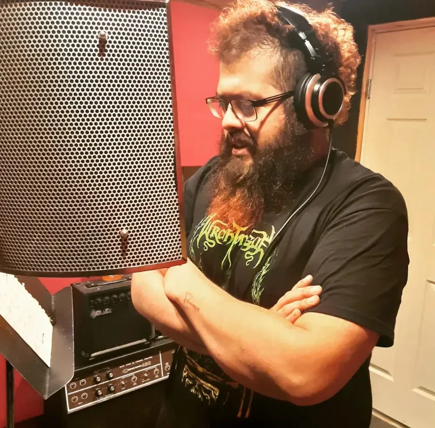 A bearded man in headphones and glasses stands with his arms crossed beside a microphone shield in a red-walled recording booth.