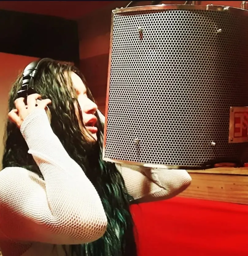 A singer with long dark green hair holds one headphone to their ear while singing into a shielded studio microphone.